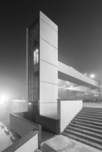 A modern, isolated urban elevator shrouded in thick fog at night captures a both mysterious and