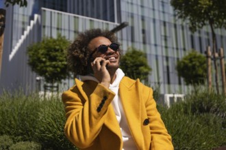 Afro man wearing a vibrant yellow coat, smiling while talking on his phone outdoors. He is seated