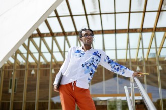 A man in a vibrant, floral shirt and orange pants poses confidently near a modern glass building,