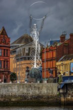 Modern metal sculpture on a riverbank in front of historic buildings under a cloudy sky, Belfast