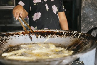 A woman prepares street food in a large wok using tongs. The image captures the sizzling oil and