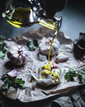 Close-up of garlic bulbs drizzled with olive oil, surrounded by fresh herbs on crumpled parchment