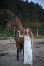 A young woman in a flowing white dress stands beside her horse on a tranquil beach. The scene