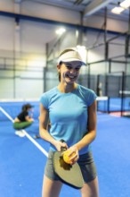 Young woman smiling and holding a pickleball paddle with a perforated ball, standing on a bright