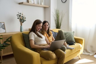 A lesbian couple sits comfortably on a yellow couch, smiling and using a laptop together in a cozy