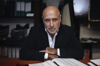 Portrait of a confident businessman seated at a desk, surrounded by office files. The scene conveys