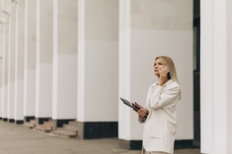A businesswoman in a beige suit talks on her phone while standing outside an office building with
