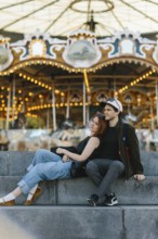 A couple sits on stone steps, the woman leaning on the man, in front of a brightly lit carousel in