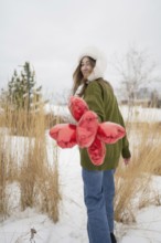 A woman wearing a green sweater and fur hat holds red heart shaped balloons in a snowy park. She
