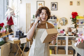 A young shop assistant wearing an apron talks on the phone while holding a cardboard box in an
