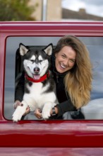 A joyful woman with long hair leans out of a red car window, holding a happy husky dog wearing a