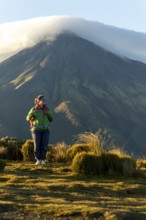 A woman wearing hiking gear stands thoughtfully in front of Mount Taranaki, New Zealand, as the