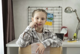 A young girl with braided hair sits on a chair in front of a white desk at home. She looks