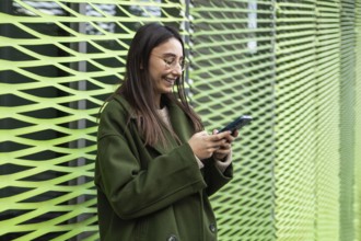 A woman wearing a stylish green coat is smiling as she uses her smartphone She stands near a