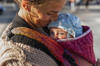 A tender moment between a mother and her baby in a colorful carrier. The warm sunlight illuminates