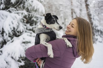 A woman in a purple jacket holds her dog lovingly in a snowy forest. Her smile and the snowy scene