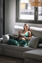 Pregnant woman in a green yoga outfit relaxes on a sofa, enjoying a healthy bowl of fruit. Sunlight