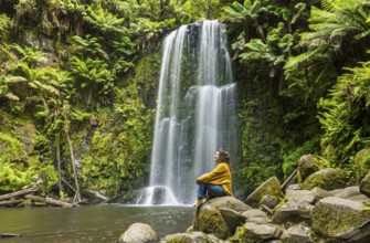 A woman sits peacefully on moss covered rocks near a waterfall surrounded by lush green ferns in