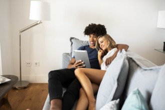A young couple relaxes on a cozy couch, sharing a tablet. The scene captures a moment of joy and