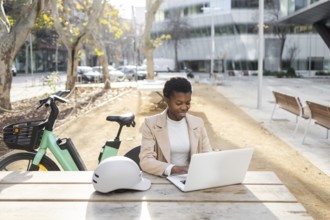 A professional black woman sits at a table in an urban park, working on her laptop Nearby, a