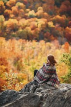A woman sits on a rocky outcrop, wrapped in a plaid blanket, savoring the vibrant hues of autumn in
