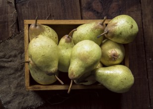 A top view of ripe pears stacked in a wooden box, placed on a dark wooden surface. The rustic