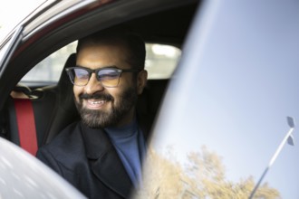 Cheerful Indian businessman looks away while smiling in his car, dressed in winter attire,