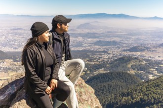 Two friends are seated on a mountain rock, gazing at a stunning panoramic view They are bundled up