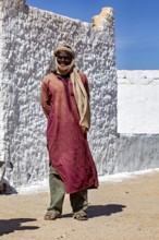 A man in traditional Tuareg clothing stands in front of a sunlit, plastered wall, Local people in