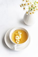 Top view of a white cup containing tea with daisies and lemon slice on a saucer. Nearby, a white