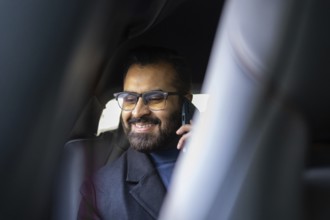A smiling Indian man dressed in a winter coat and sweater, talking on a mobile phone while sitting