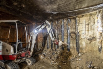 An excavator operates in an underground construction site, tearing through rock and earth. The dim