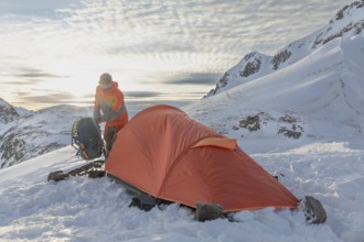 A ski tourer sets up near a bright orange tent amidst snowy mountains at sunrise, capturing the