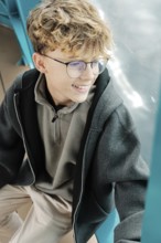 A curly-haired teenager with glasses sits by a window, smiling as he enjoys a ski resort in the