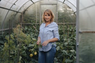 A woman in casual attire stands amidst lush plants inside a greenhouse. She tends to the greenery,