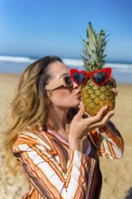 Woman playing with a pineapple wearing red heart sunglasses, standing on a sandy beach by the