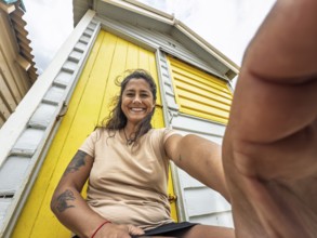 A joyful woman takes a close-up selfie in front of a vibrant yellow Brighton bathing box in