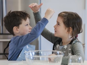 Two young siblings wearing star patterned aprons are joyfully interacting in a kitchen setting. A