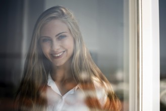 A young woman smiles gently through a glass window, captured in soft natural light. Her relaxed