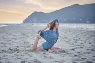 A woman does Eka Pada Rajakapotasana on a sandy beach while the sun sets over the water and hills