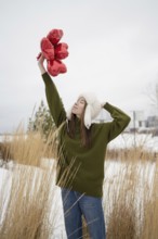 A young woman in a green sweater and white hat lifts red heart shaped balloons in a snowy park. The