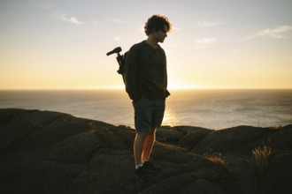 A young male photographer stands on the rocky terrain of Cape Spear Lighthouse, Newfoundland &