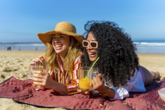 Two cheerful young women lying on a red blanket at the beach, laughing and enjoying their drinks