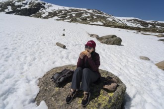 An Asian woman sits comfortably on a rocky surface, enjoying a meal amidst snow-covered peaks in