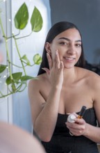 A woman stands by a mirror, applying skincare cream with a smile. Natural light and indoor plants