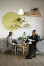 Two women enjoy a friendly chat over coffee in a cozy, stylish cafe with modern decor and lush
