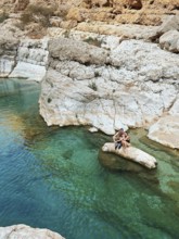 A young smiling couple relaxes on a rocky outcrop in the emerald waters of Wadi Ash Shab, a