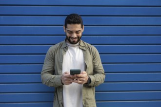 A man in a green shirt and white T shirt smiles while using his smartphone in the city. He stands