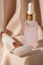 A sleek jade roller and a glass dropper bottle containing pink serum sit on elegant fabric These