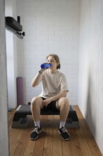 A young person takes a refreshing break with a water bottle in a compact home gym, sitting on a